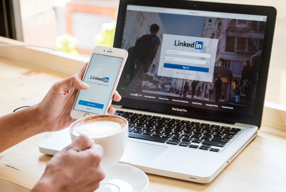 A marketer holds a smartphone and a coffee in front of a laptop (for some reason). Both devices display LinkedIn login pages.