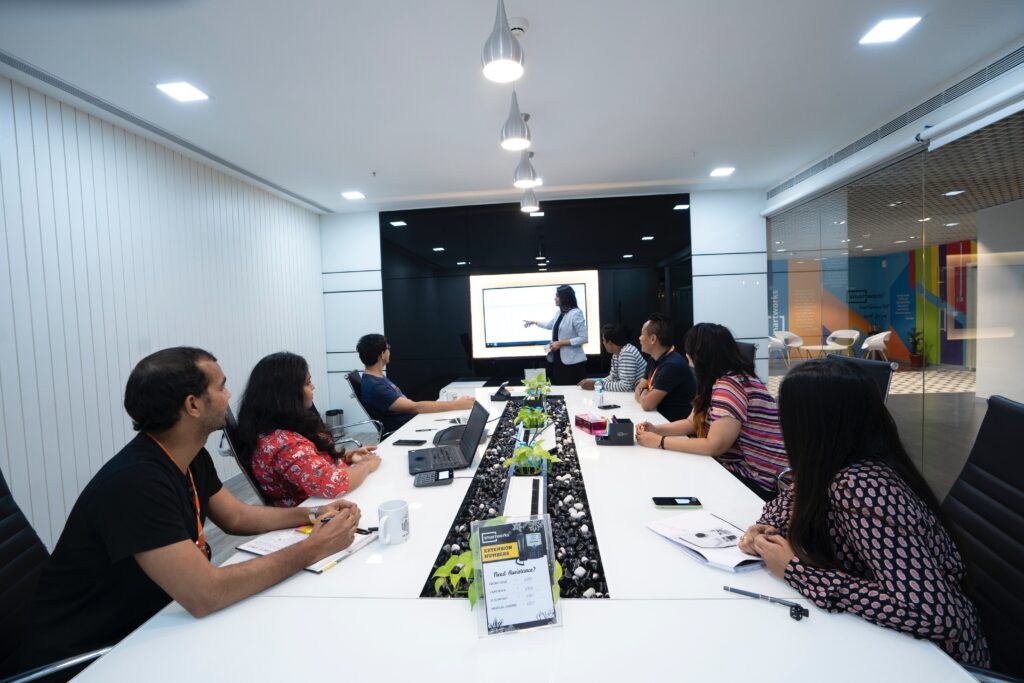 A group of office workers seated at a large conference table watch a presentation on screen