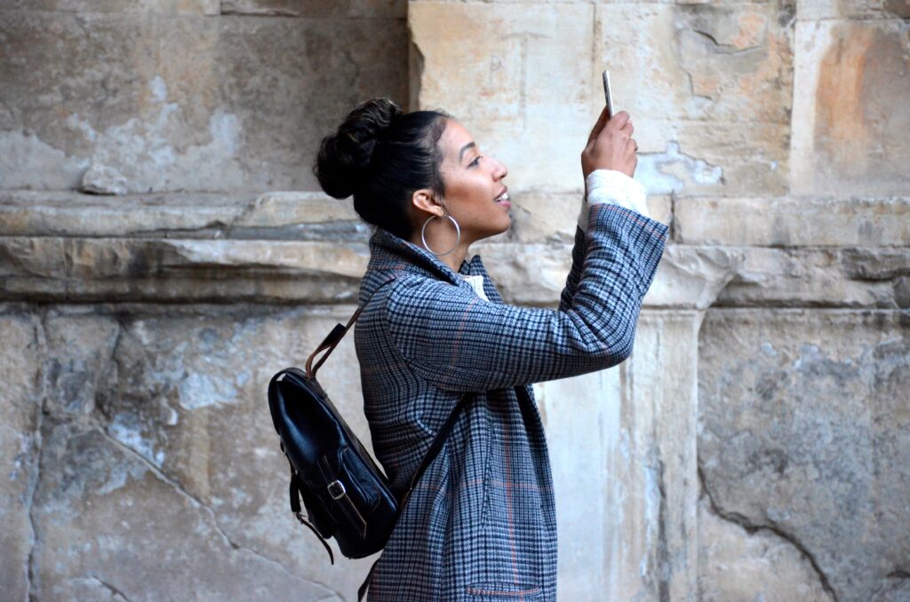 A well-dressed woman social media manager uses a smartphone to take a picture outside with a weathered stone wall in the background.
