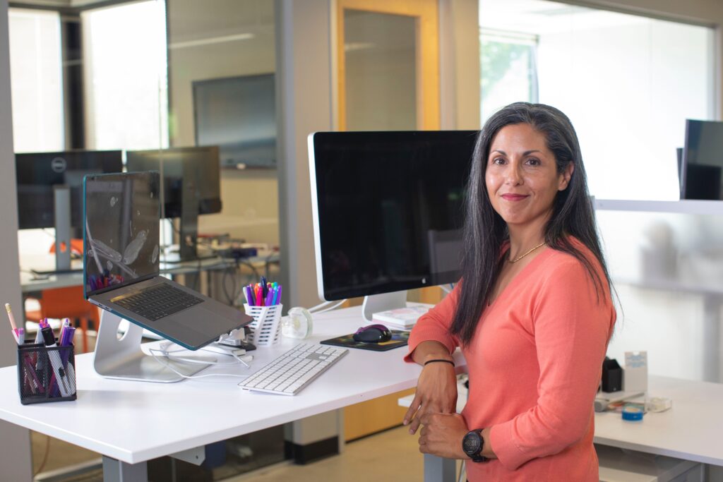 A professional woman representing a buyer's persona stands at a desk with laptop and monitor at the ready, looking confidently to camera