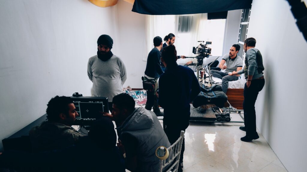 Wide shot of a video production in a small white room. Various crewmembers set up their work, from production staff in the foreground, to actors and camera operators in the background.