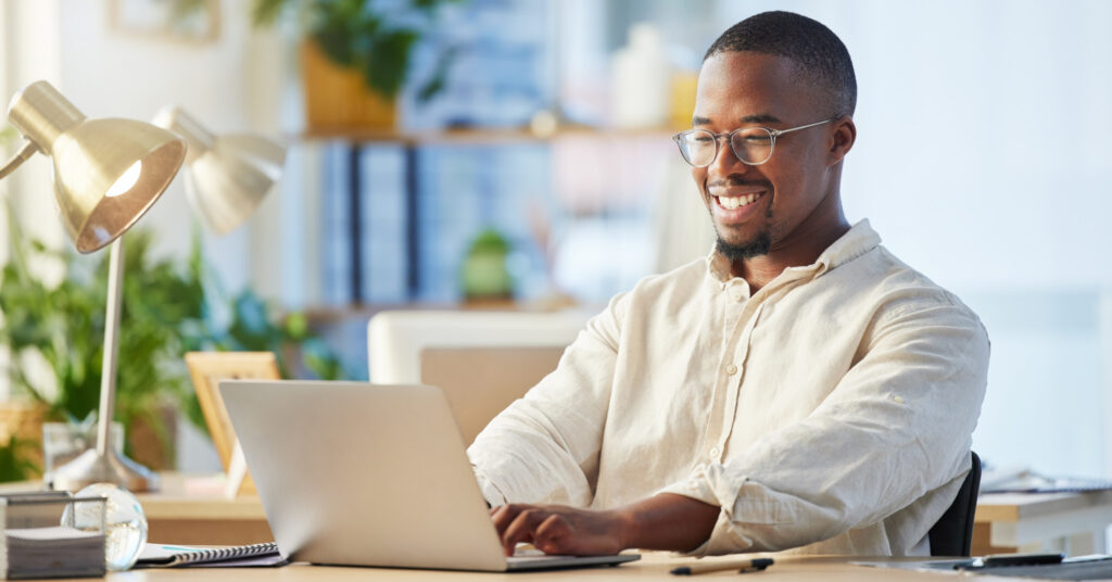 A black professional in an office types on a laptop while smiling at the great email subject line he's writing.