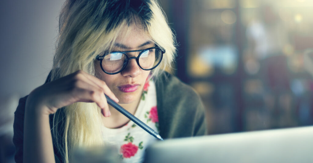 A young woman with a pen looks at a laptop and ponders her SEO FAQs