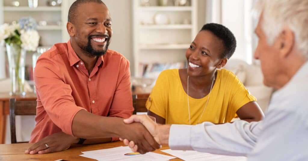 A married couple shakes hands with their wealth management professional across a kitchen table thanks to marketing.