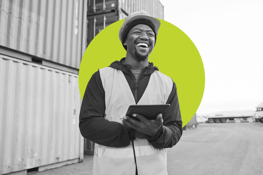 A worker in safety vest and hardhat stands in front of shipping containers and laughs, a tablet in hand. 