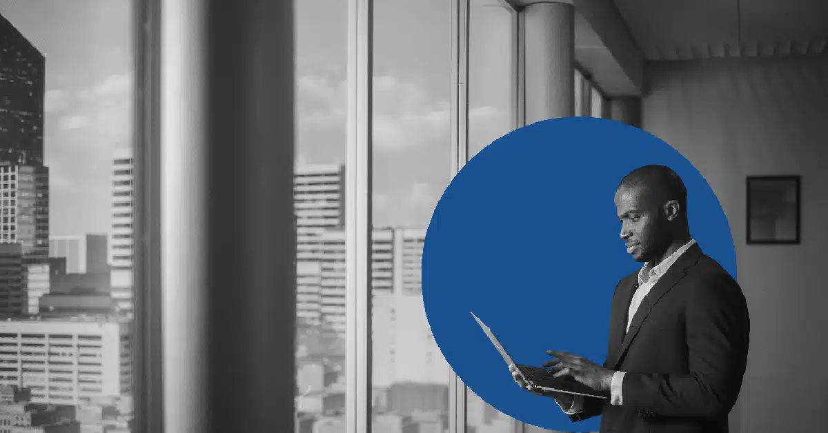 a man looks out a window at a city skyline while checking email on a laptop