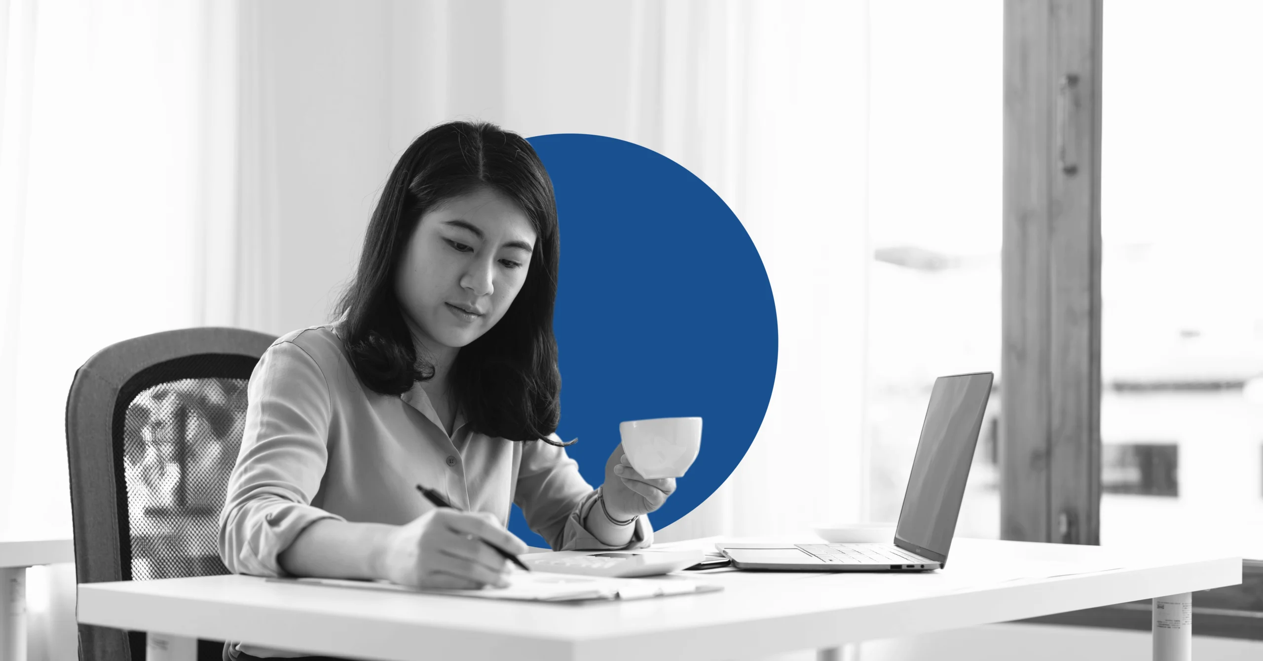 a businesswoman drinks from a cup and takes notes while working on a laptop